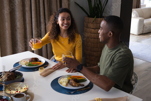 Diverse couple sitting at table in mustard top, holding orange juice and roasted chicken plates