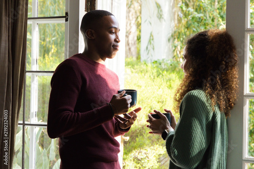 Diverse couple standing by French door, holding mugs, talking with smartwatch visible