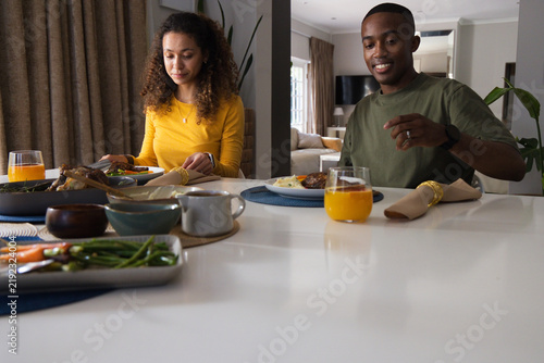 Diverse couple sitting at home table, woman wearing yellow, man in olive tee reaching orange juice