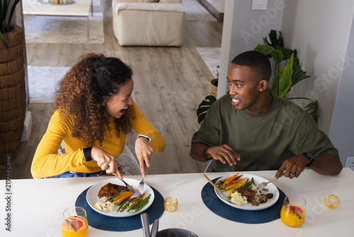 Couple cutting and talking at home dining table, woman mustard top, man olive tee, orange juice