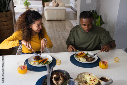 Couple wearing yellow top and olive t-shirt cutting meal at home dining table with orange juice