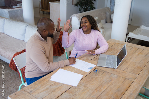 African American couple sitting at home dining table, signing paperwork with laptop, blue pen, keys