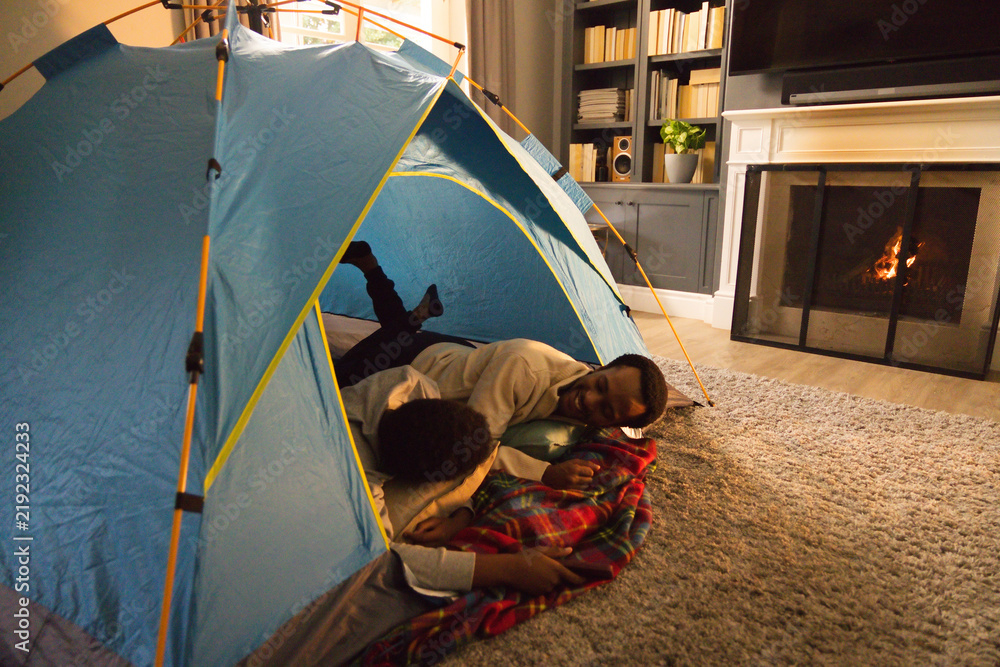Obraz premium African American father and child lying in blue tent on gray rug in living room, talking