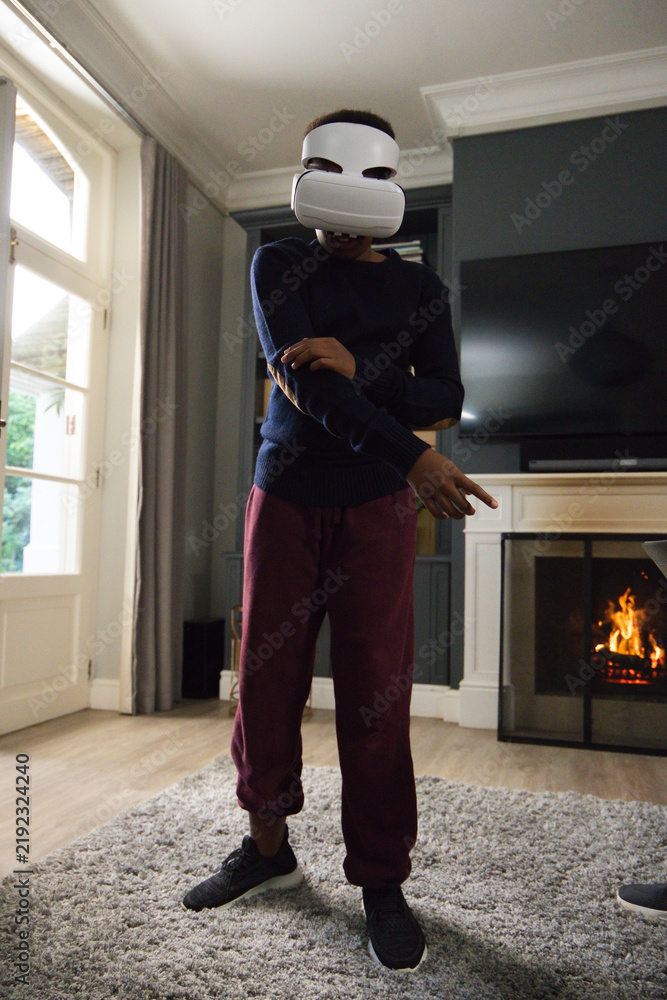 Obraz premium African male youth wearing white virtual reality headset, standing on rug in living room near TV