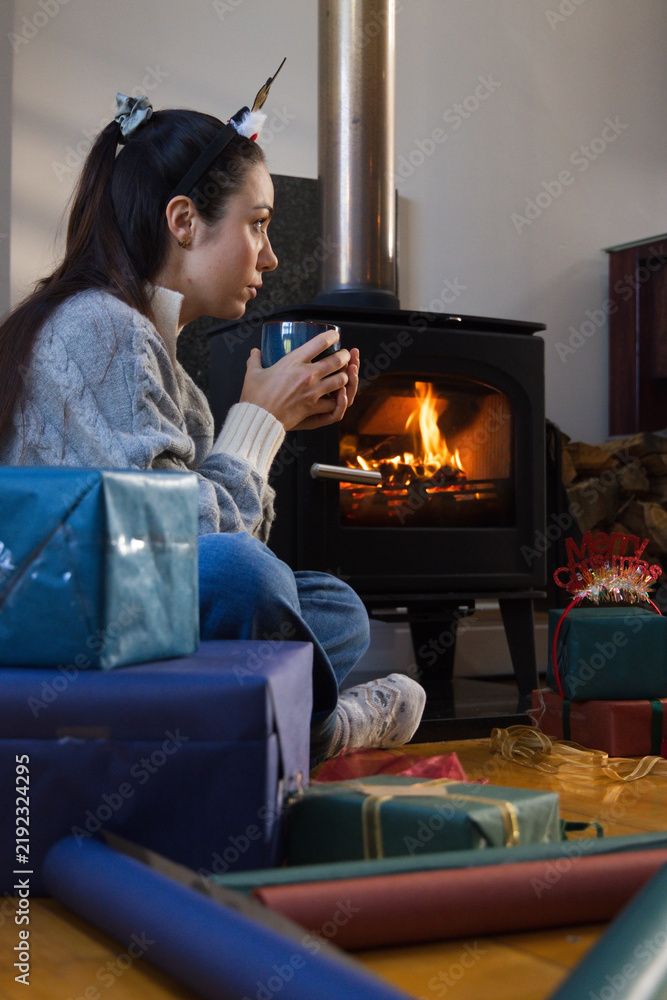 Obraz premium Woman wearing knit sweater holding mug sitting by wood stove with wrapped gifts and paper