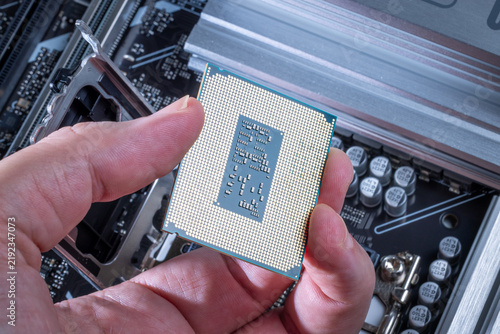 CPU with LGA socket held in technician's hand. Background shows motherboard during PC assembly or upgrade. Visible motherboard components. High-end computer parts