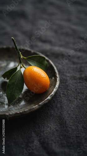 A fresh whole kumquat with leafy twig on dark ceramic dish over linen