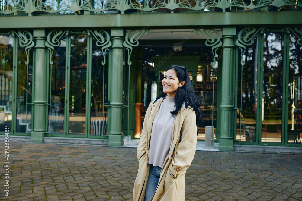 Obraz premium Woman in a beige coat outdoors, smiling beside a green decorative entrance, park backdrop, casual fashion vibe, candid mood, daylight and greenery