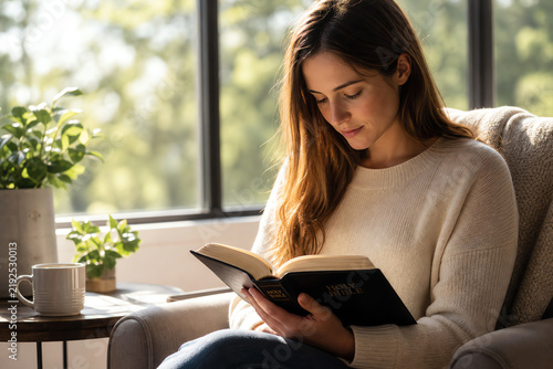 Young woman reads Holy Bible in cozy armchair by sunny window with plants and coffee, depicting peaceful faith