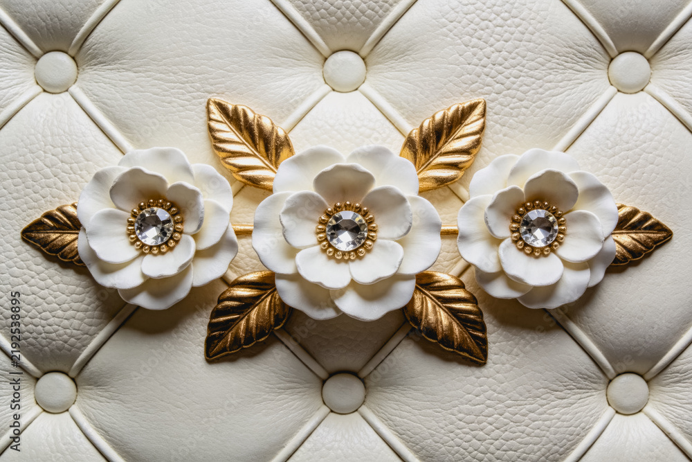 Fototapeta premium Three white floral decorations with gold leaves and central gems sit on a quilted light cream leather surface, creating an elegant decorative detail.