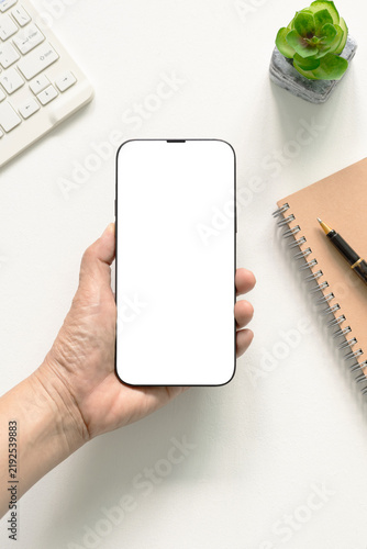 Top view flat lay hand holding modern smartphone with blank white screen template, keyboard, notebook, pen, and plant on white desk. Concept for marketing, website, branding, social.