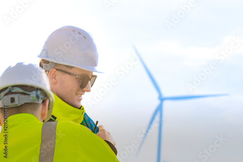 A fair-haired technician in dark sunglasses looks out across the field with a blurred turbine floating over his shoulder, hazy evening light softening the whole frame