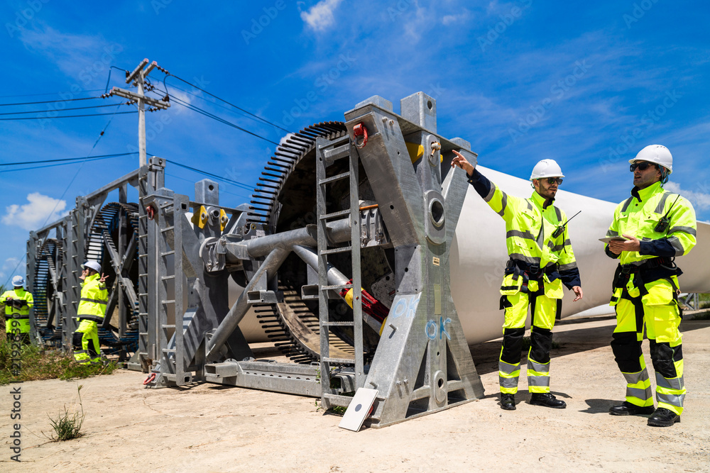 Fototapeta premium An engineer points up at the pitch bearing teeth of a wind turbine hub as his colleague makes notes on a tablet beside him at a green energy construction site in tropical daylight.