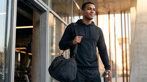 Medium shot of happy man carrying duffel bag and stainless steel water bottle walking out of modern gym entrance, post workout concept.