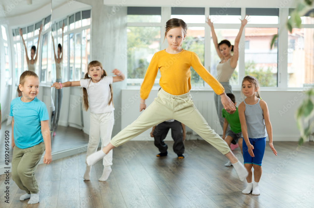 Fototapeta premium Girl engaged jumping into air in fitness studio with friends, group of youthful friends bob up and down during dance class. Children in good mood actively move and dance at choreography.