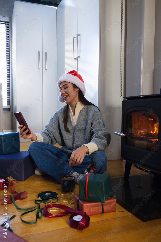 Obraz premium Adult female sitting on floor near stove, wearing Santa hat, holding smartphone, wrapping gifts