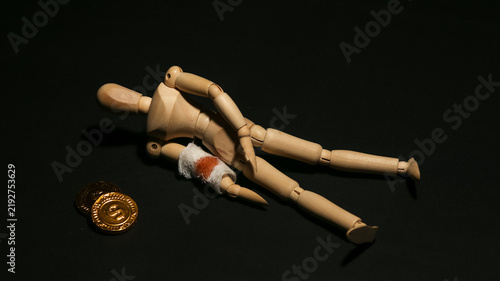 Fotografie Injured wooden mannequin lying down with a bloody bandage on its arm next to gold coins, symbolizing the cost of greed, sacrifice, or financial loss