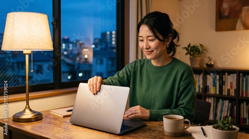 Asian woman closing laptop at desk in cozy home office at night.