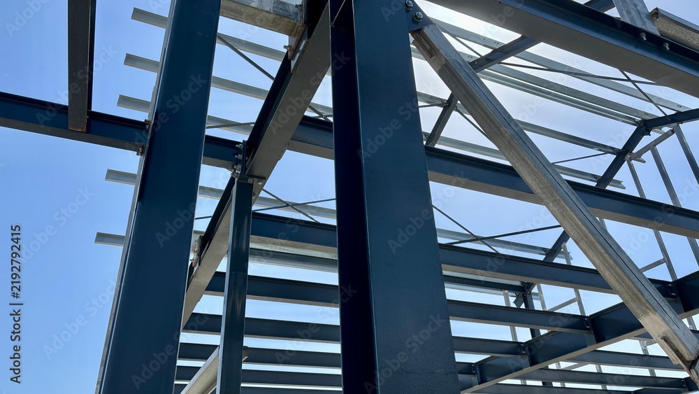 Obraz premium Unfinished steel building frame with blue structural beams, metal roof supports and a wooden ladder against a clear sky at a construction site