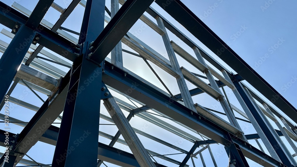 Obraz premium Steel structural framework of an unfinished building with blue beams, galvanized metal supports and bolted connections against a blue sky