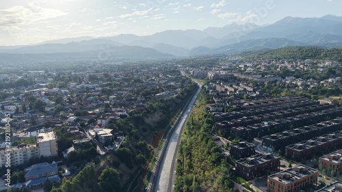 Aerial flyover above river park promenade toward mountains with luxury residential complexes at sunrise