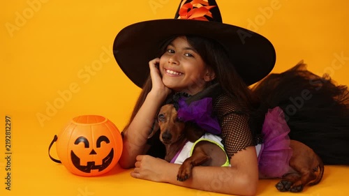 little brunette girl in a witch costume for Halloween against a yellow background in a studio with a dachshund dog, space for text