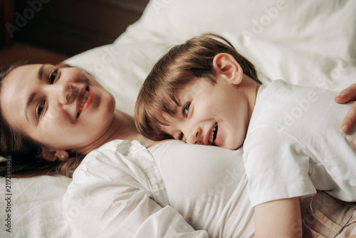 A happy mother and her young son are lying in an embrace on the bed.