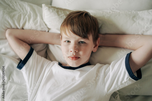 Young boy lying on bed with hands behind his head looking relaxed in bright bedroom