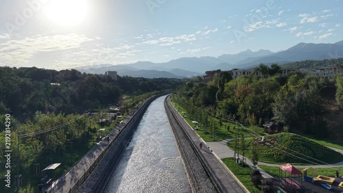 Aerial flyover above river park promenade toward mountains with luxury residential complexes at sunrise
