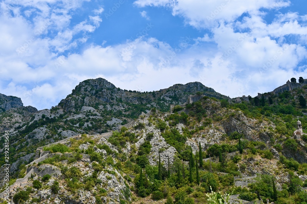 Fototapeta premium A scenic horizontal view of medieval stone fortifications stretching along steep mountain ridges, with a church belfry visible in the lower foreground under a partly cloudy blue sky.