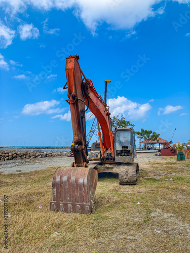 Принти на полотні An orange excavator parked on a coastal field under a bright blue cloudy sky