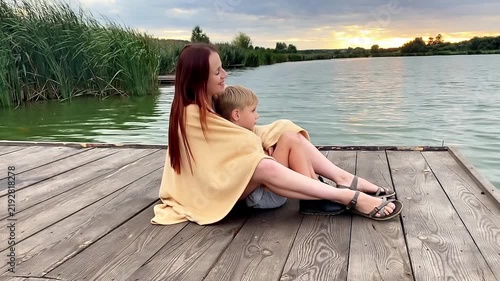 Woman and little child boy sitting near river.Happy family mother kid talking,having fun,hugging in nature against lake.Parenting,child care.Healthy baby time outdoors