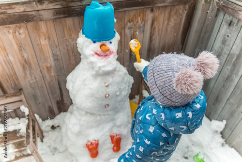 Obraz na płótnie Little sister feeding handmade snowman with spoon in backyard, funny winter game