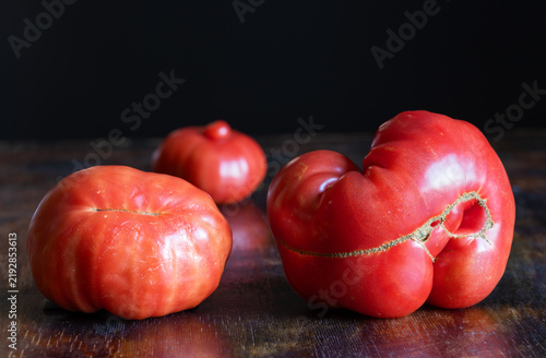 Three unusual, naturally misshapen red tomatoes on rustic wooden table. Tomatoes have pronounced irregular shapes, deep grooves, scars, surface cracking. Food that does not meet standard expectations