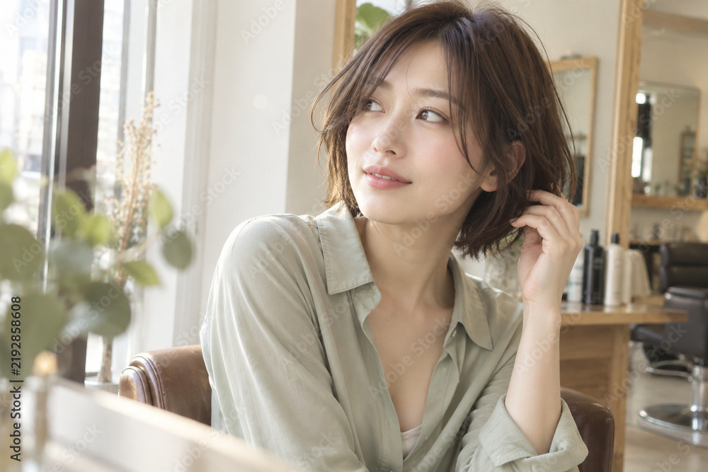 Obraz premium 女性が淡い緑のシャツ姿で毛先に触れ美容室の窓の方を見る