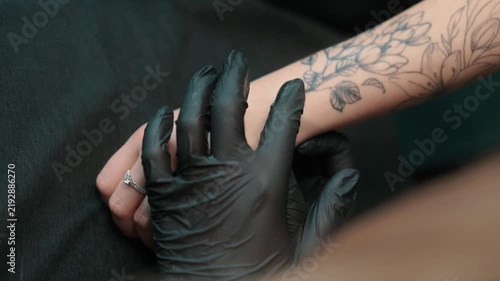 A tattoo artist in black gloves positions a floral stencil on a clients tattooed forearm during a closeup preparation process.