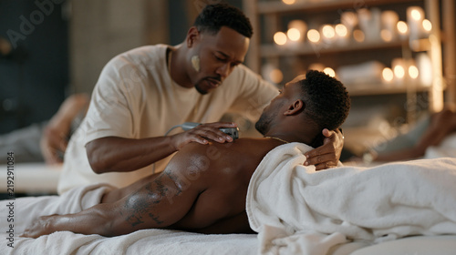 A boxing trainer in a candlelit recovery gym tests a new muscle percussion tool on a training partner's shoulder, a smudge of liniment on the trainer's cheek, the diverse team of