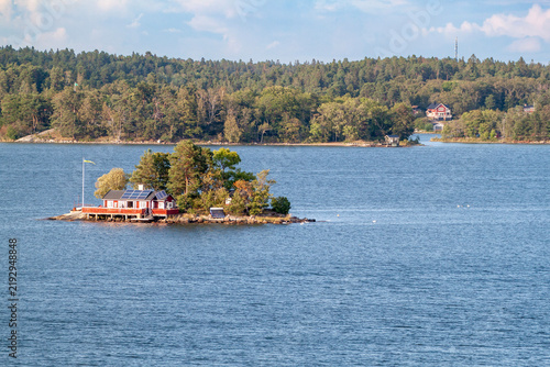 Small isolated island with red wooden cottage in Stockholm archipelago. Traditional swedish summer house on tiny islet surrounded by sea water and green forest background.