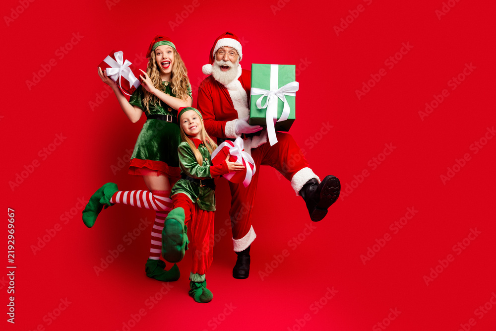 Obraz premium Santa Claus and two cheerful elves presenting wrapped gifts and celebrating joyful Christmas family fun with laughing adults and child on a vibrant red background