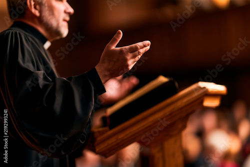 Man priest standing at wooden pulpit giving sermon to church congregation. Speaker gesturing while preaching to audience in religious place. Faith, spirituality and community gathering
