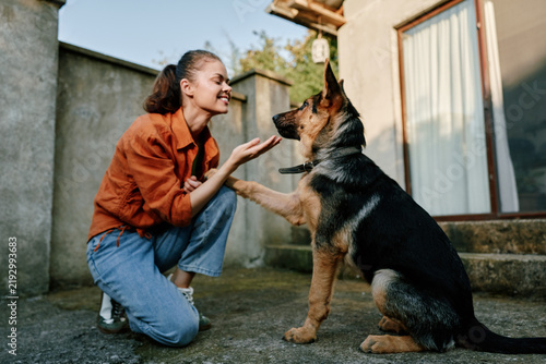 Принти на полотні A cheerful woman kneels on a concrete yard, offering a high five to a dog as they share a warm, playful moment