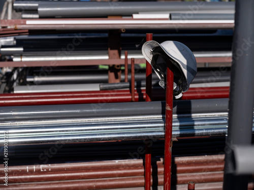 Konstfotografi A white safety helmet hangs on a red post beside racks of stainless steel and coated pipe at a shipyard