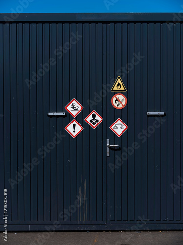 Ghs hazard pictograms and a no smoking sign mark the dark doors of a chemical storage unit at a shipyard.