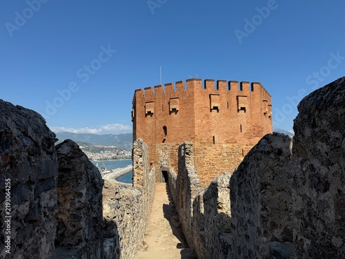 Foto Scenic view of the historic Red Tower (Kızılkule) from inside the ancient fortress walls in Alanya, Turkey, featuring a blue sky, stone walkway, and coastal harbor background