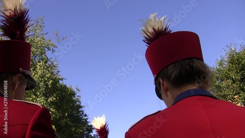 Back side of heads of two women brass orchestra musicians wearing red uniform hats decorated with feathers against blue sky background.