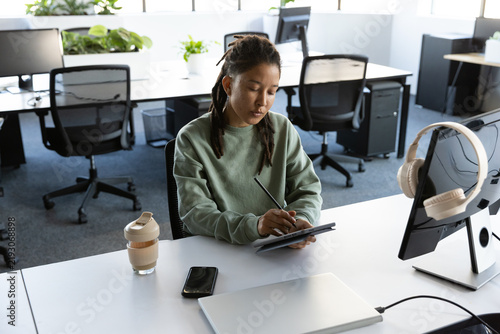 Nonbinary African American wearing green sweater sitting at open office desk using stylus on tablet