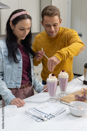 Couple preparing pink milkshakes in parfait glasses at kitchen countertop, adding cherries
