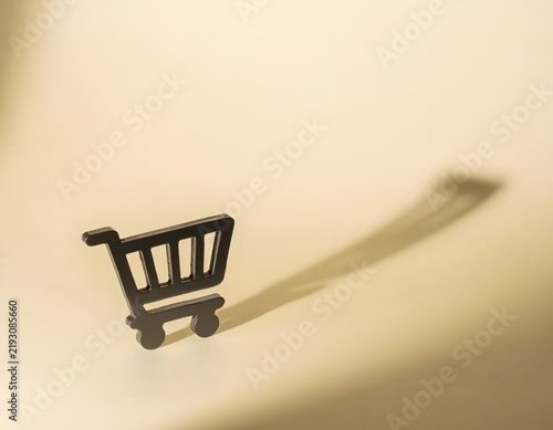 Small Black Shopping Cart Casting Large Dramatic Shadow on Cream Background, Minimal Ecommerce and Retail Concept