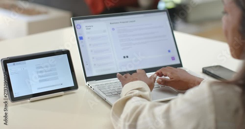 Hands are reaching and tapping laptop keyboard at desk, are typing to respond to tablet chat