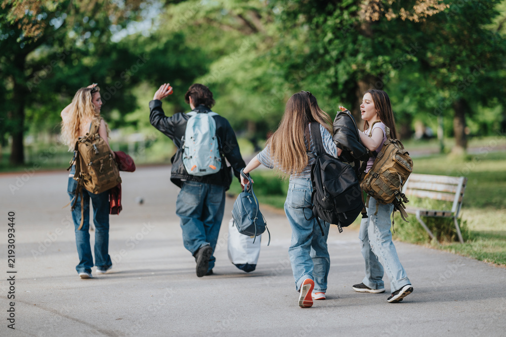 Obraz premium A group of teen friends walking together carrying backpacks along a tree-lined park path. Four teenagers chat and laugh while strolling through green outdoor surroundings.